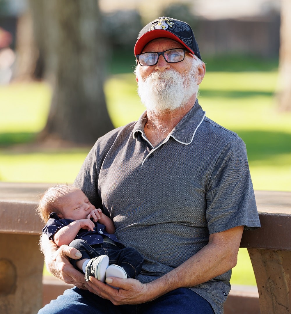 William Metcalf, grateful patient, holding his grandchild while setting at a picnic table in a park.